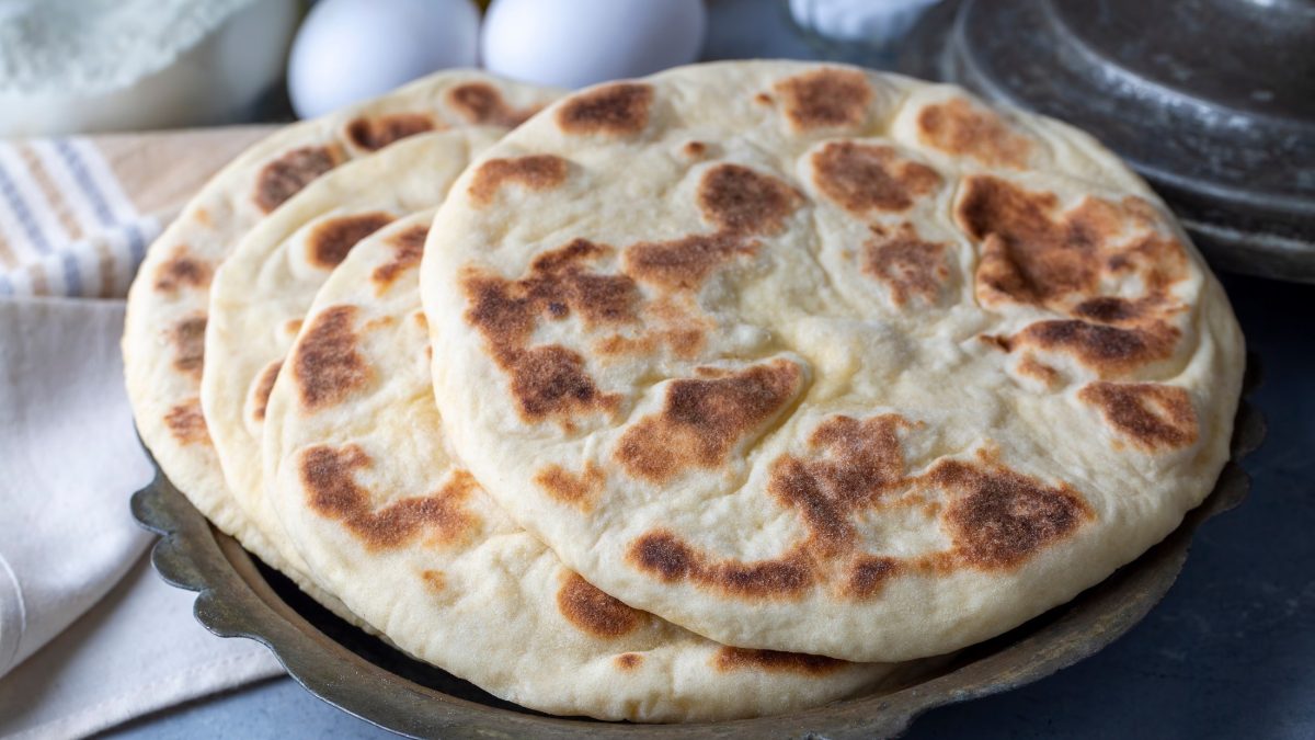 Draufsicht, schräg: Zwei Bazlama, also türkische Fladenbrote, liegen auf einem rustikalen Tisch. Daneben liegen zwei Handtücher und im Hintergrund drei Eier.