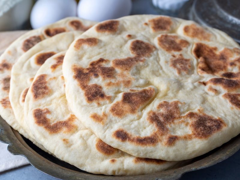 Draufsicht, schräg: Zwei Bazlama, also türkische Fladenbrote, liegen auf einem rustikalen Tisch. Daneben liegen zwei Handtücher und im Hintergrund drei Eier.