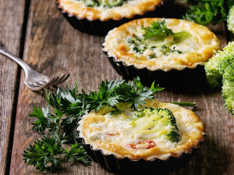 Draufsicht, schräg: Drei Brokkoli-Quiche in kleinen Tarte-Formen auf einem rustikalen Holztisch.