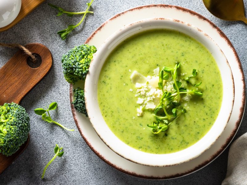 Draufsicht: Auf einem bläulichem Untergund steht eine runde Schale auf einem Teller. In der Schüssel ist eine Brokkoli-Suppe mit Kartoffeln. Drum herum liegen Brokkoli-Röschen und Brokkoli-Sprossen sowie ein Löffel.