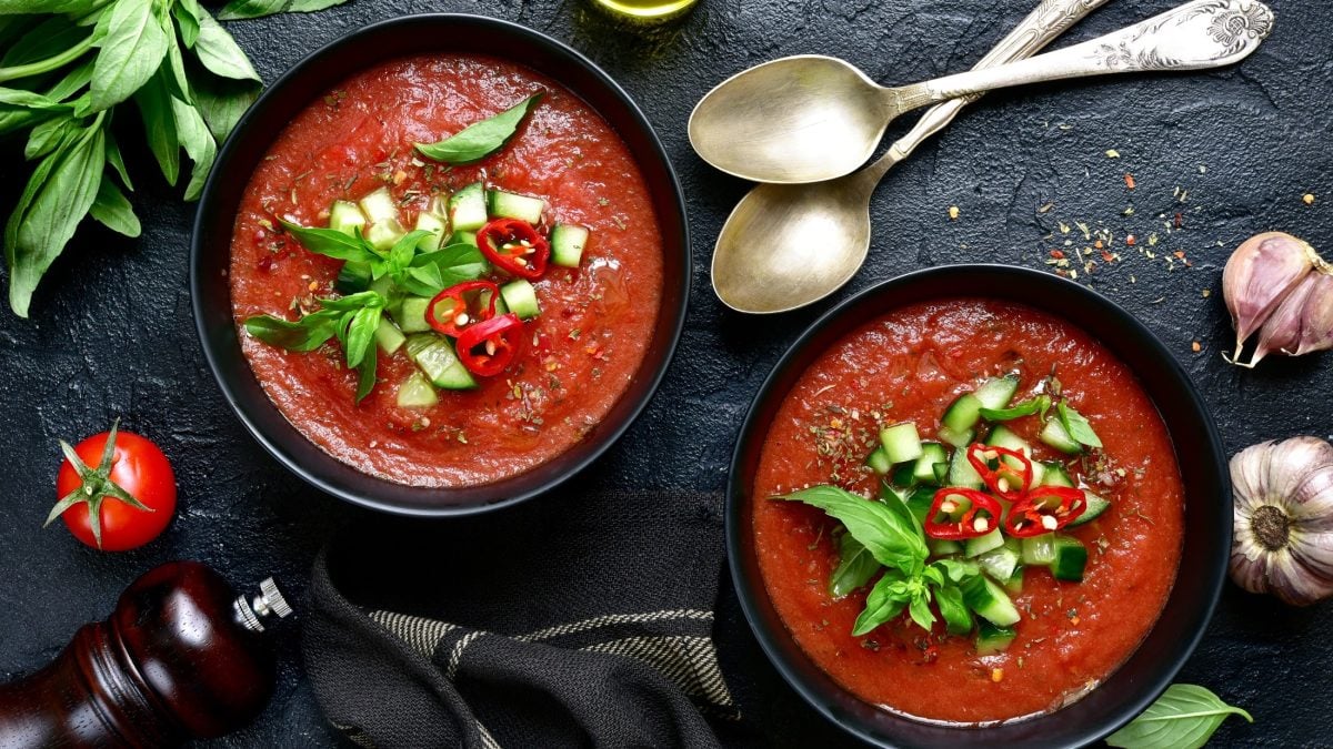 Draufsicht: Zwei Schüssel mit einer Gazpacho Andaluz auf einem dunklen, rustikalem Untergrund. Drum herum liegen frische Kräuter, Gewürze und zwei silberne Suppenlöffel.