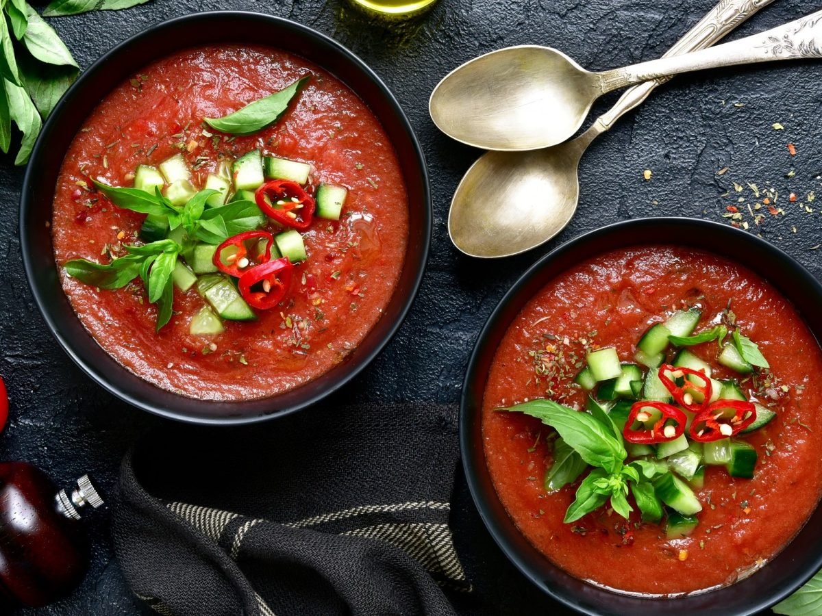 Draufsicht: Zwei Schüssel mit einer Gazpacho Andaluz auf einem dunklen, rustikalem Untergrund. Drum herum liegen frische Kräuter, Gewürze und zwei silberne Suppenlöffel.