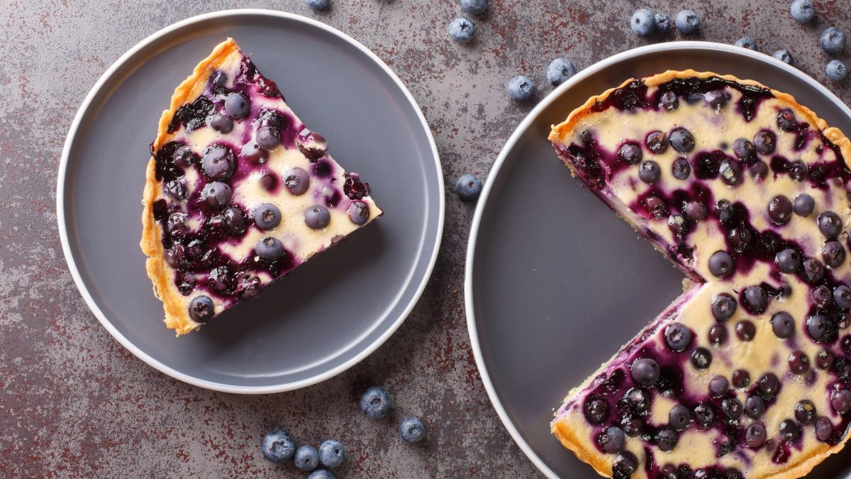 Draufsicht: Ein runder angeschnittener Käsekuchen mit Heidelbeeren. Daneben liegt ein weiterer Teller mit einem Stück Käsekuchen. Drum herum liegen frische Heidelbeeren.