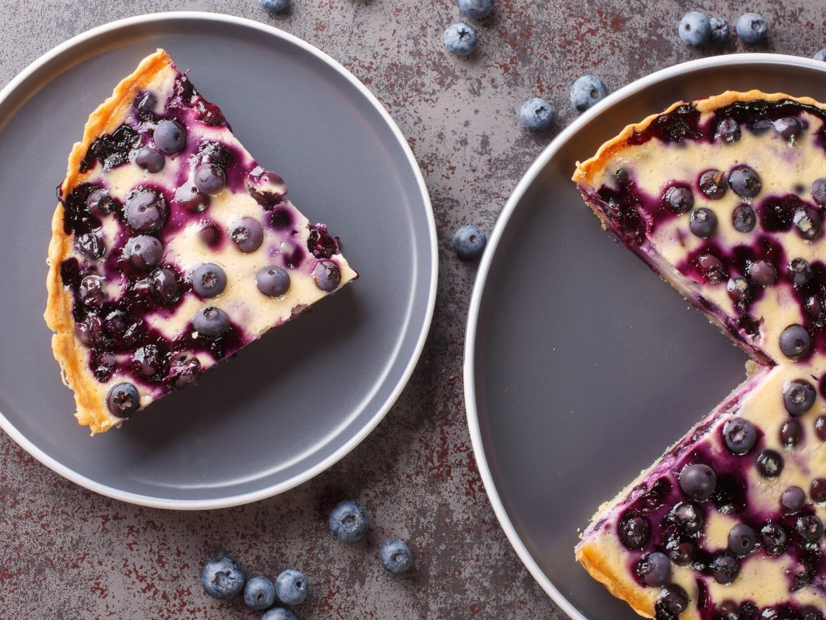 Draufsicht: Ein runder angeschnittener Käsekuchen mit Heidelbeeren. Daneben liegt ein weiterer Teller mit einem Stück Käsekuchen. Drum herum liegen frische Heidelbeeren.