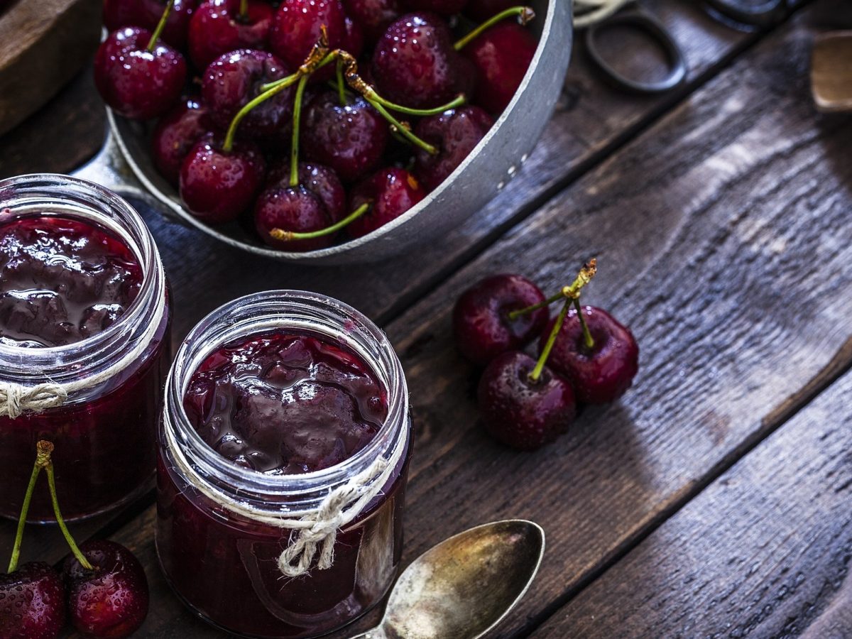 Draufsicht: Zwei Gläser Kirsch-Balsamico-Marmelade auf einem dunklen, rustikalen Holztisch. Daneben liegen ein Sieb mit frischen Kirschen, sowie einzelnen Kirschen.