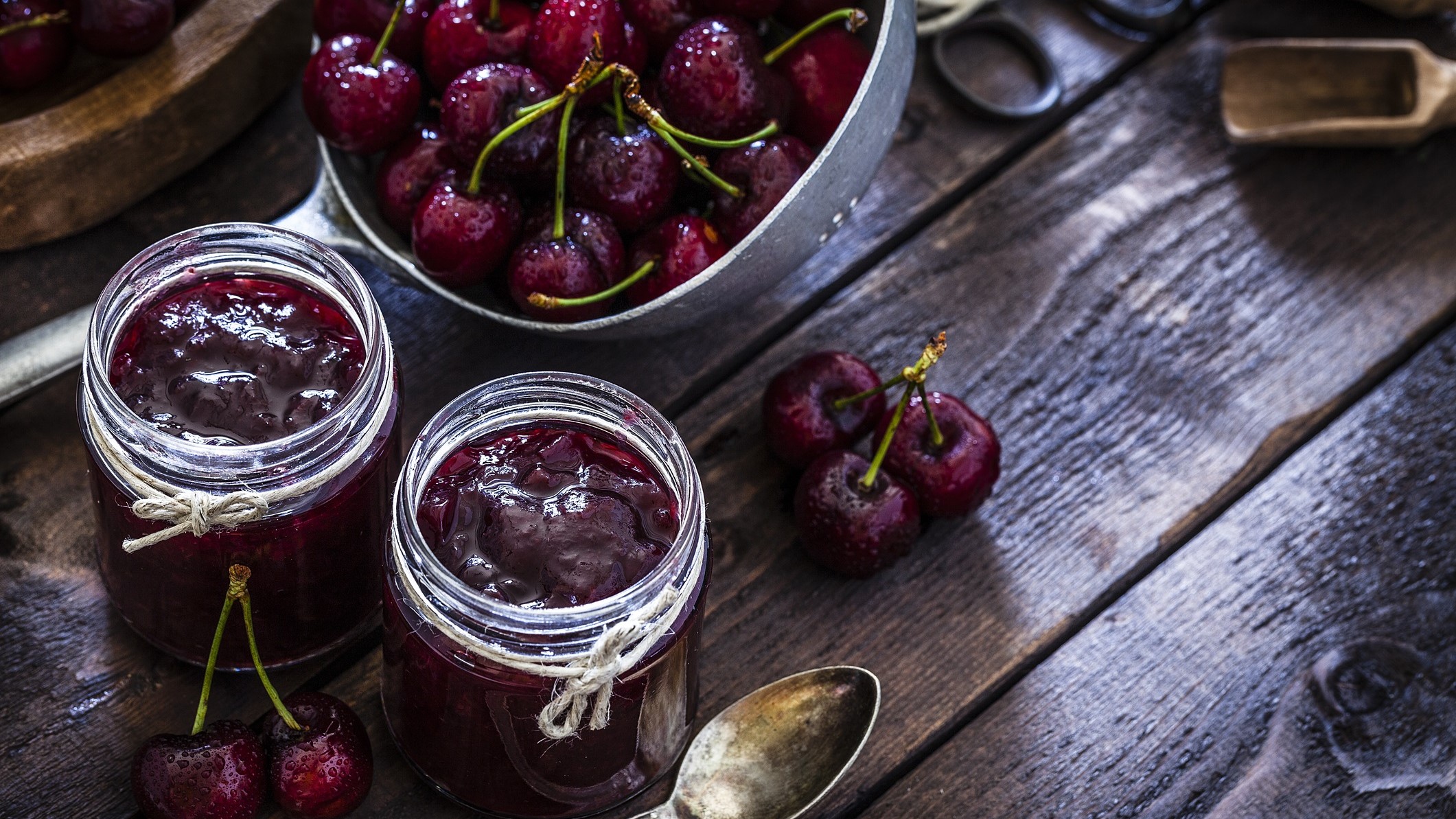 Draufsicht: Zwei Gläser Kirsch-Balsamico-Marmelade auf einem dunklen, rustikalen Holztisch. Daneben liegen ein Sieb mit frischen Kirschen, sowie einzelnen Kirschen.