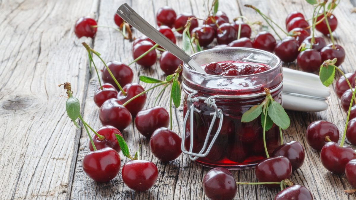 Frontal: Ein geöffneten Glas Kirsch-Chutney steht auf einem rustikalen Holzboden aus Dielen. Drum herum liegen viele frische Kirschen.