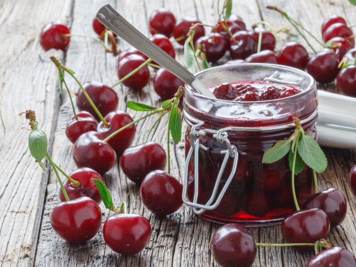 Frontal: Ein geöffneten Glas Kirsch-Chutney steht auf einem rustikalen Holzboden aus Dielen. Drum herum liegen viele frische Kirschen.