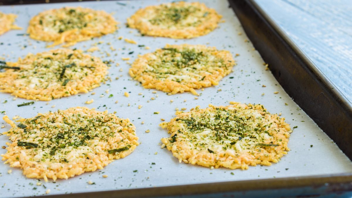 Draufsicht schräg: Ein Backblech mit selbst gemachten Parmesan-Pistazien-Chips auf einem hellen, blauen Untergrund.