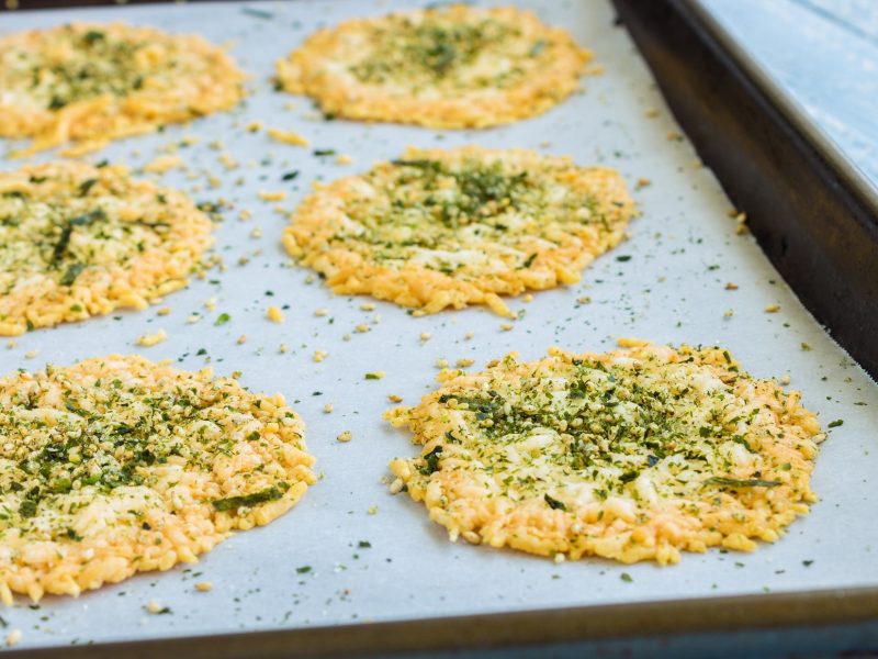 Draufsicht schräg: Ein Backblech mit selbst gemachten Parmesan-Pistazien-Chips auf einem hellen, blauen Untergrund.