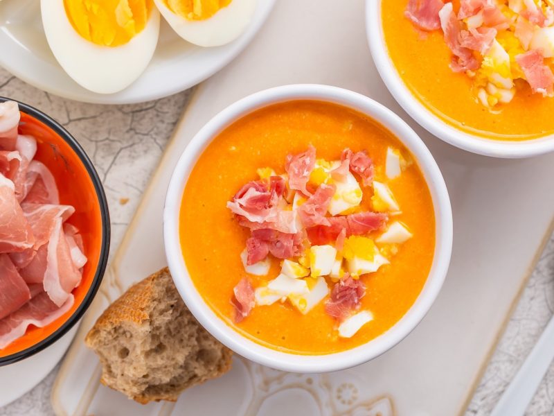 Draufsicht: Ein weißer Untergrund aus Marmor mit zwei weißen Schalen. Die sind Salmorejo andaluz gefüllt. Das ist eine spanische kalte Tomatensuppe. Drum herum sind zwei Schalen: Eine mit Schinken und eine mit hart gekochten Eiern.