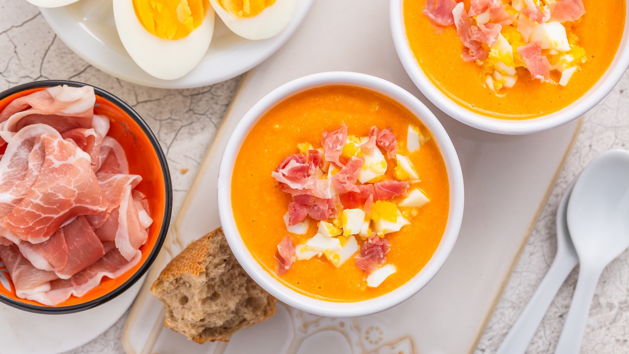 Draufsicht: Ein weißer Untergrund aus Marmor mit zwei weißen Schalen. Die sind Salmorejo andaluz gefüllt. Das ist eine spanische kalte Tomatensuppe. Drum herum sind zwei Schalen: Eine mit Schinken und eine mit hart gekochten Eiern.