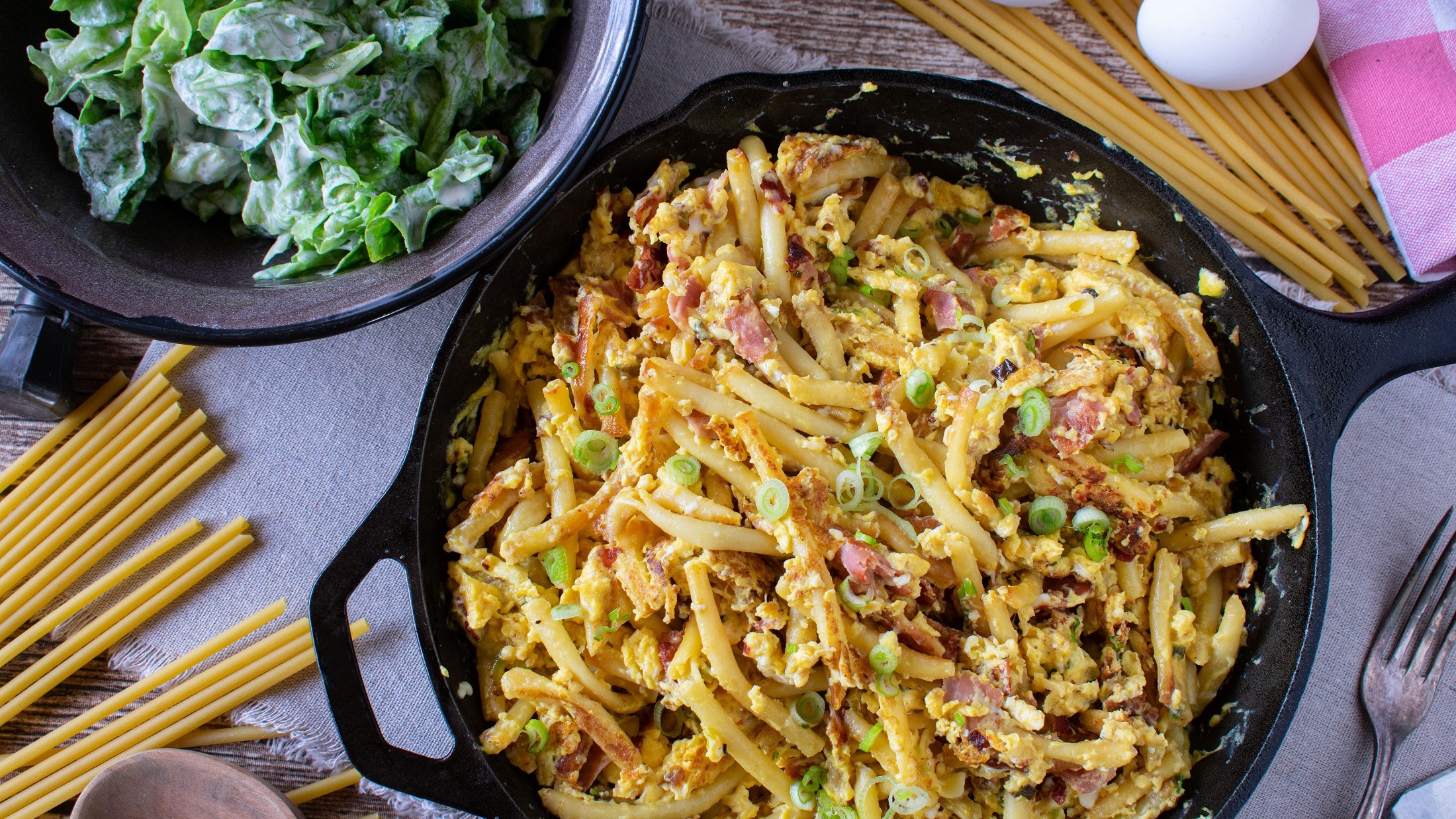 Draufsicht: Eine Pfanne Schinken-Nudeln mit Frühlingszwiebeln. Drum herum liegen rohe Nudeln und ein rohes Ei. Daneben steht eine Schüssel mit einem frischen grünen Salat.