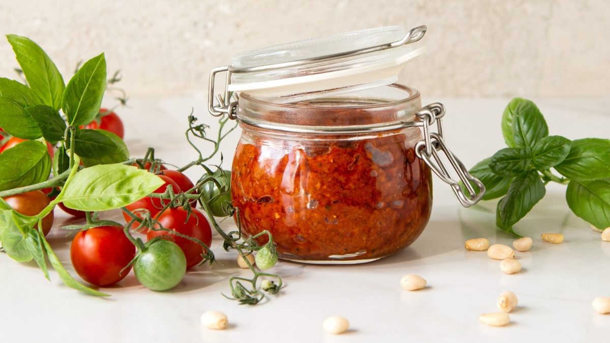 Frontal: Weißer Untergrund, ein Weck-Glas mit Tomaten-Pinienkern-Pesto. Drum herum liegen frische rote und einige grüne Tomaten, Pinienkerne und Basilikumblätter.