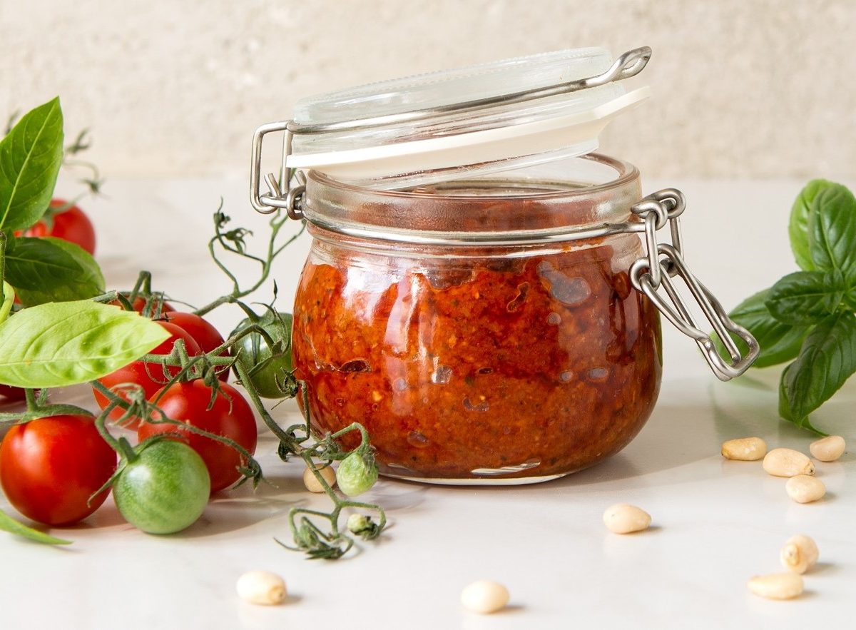 Frontal: Weißer Untergrund, ein Weck-Glas mit Tomaten-Pinienkern-Pesto. Drum herum liegen frische rote und einige grüne Tomaten, Pinienkerne und Basilikumblätter.