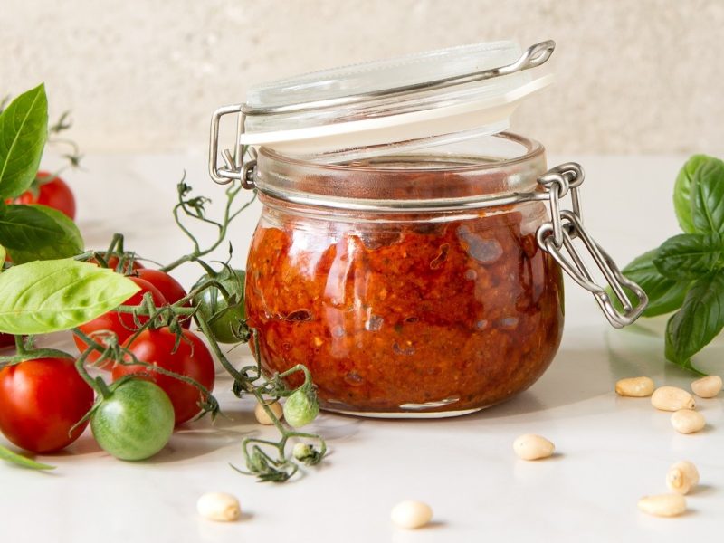 Frontal: Weißer Untergrund, ein Weck-Glas mit Tomaten-Pinienkern-Pesto. Drum herum liegen frische rote und einige grüne Tomaten, Pinienkerne und Basilikumblätter.