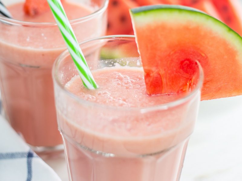 Frontal: Zwei Gläser mit einem Wassermelonen-Smoothie mit je einem Strohhalm. Die Gläser sind mit einem Scheibe Wassermelone dekoriert.