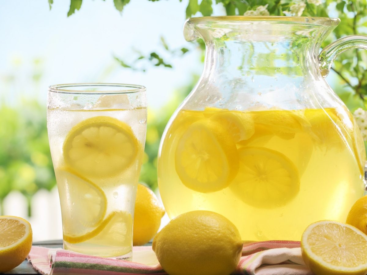 Frontalsicht: Garten, Sommer. Auf einem Tisch stehen ein Glas und ein Krug mit Zitronenlimonade. Drum herum liegen Zitronenhälften. Zitronenlimonade selber machen.