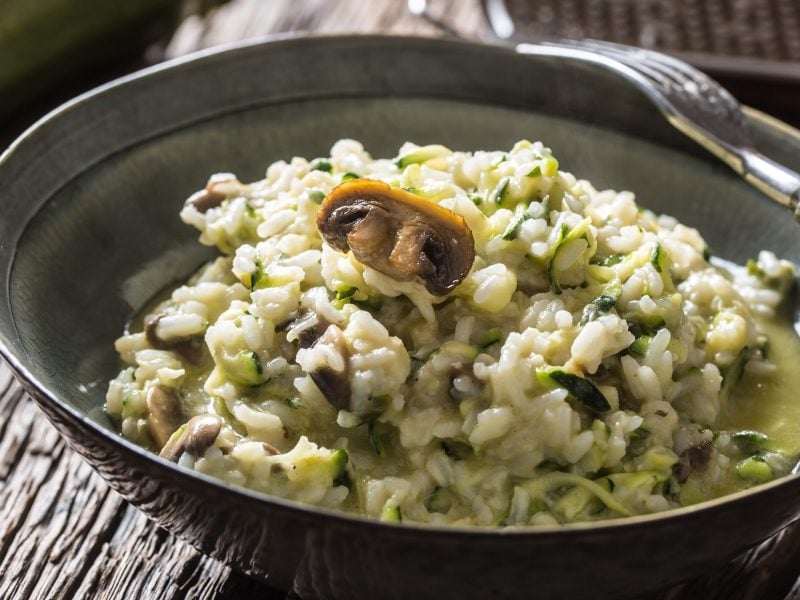 Draufsicht, schräg: Eine weiße Schüssel Zucchini-Risotto mit Pilzen auf einem rustikalen Untergrund aus Holz.