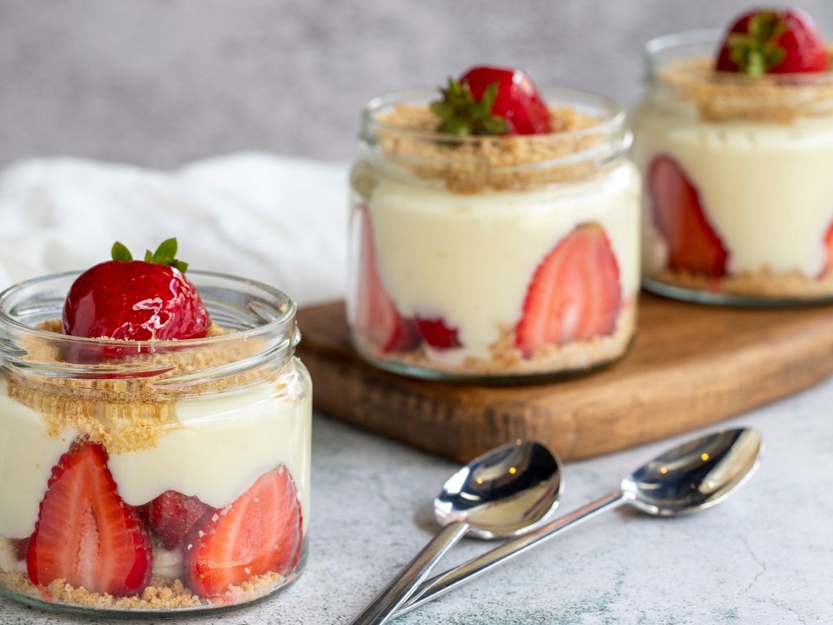 Drei Gläser mit dem Dessert auf einem grauen Tisch mit kleinem Holzbrett. Alles von der Seite fotografiert.