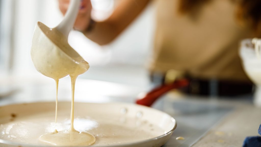 Teig aus Grundrezept für Pfannkuchen wird von einer Frau mit einer weißen Kelle in eine helle Pfanne gegeben.