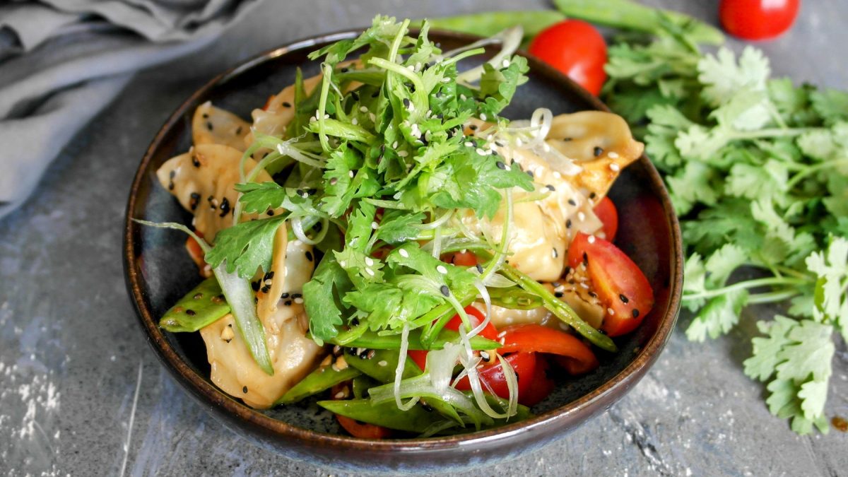 Eine schwarze Schüssel mitd em Gyoza-Nudelsalat udn Kräutern auf einem grauen Steintisch von schräg oben fotografiert.