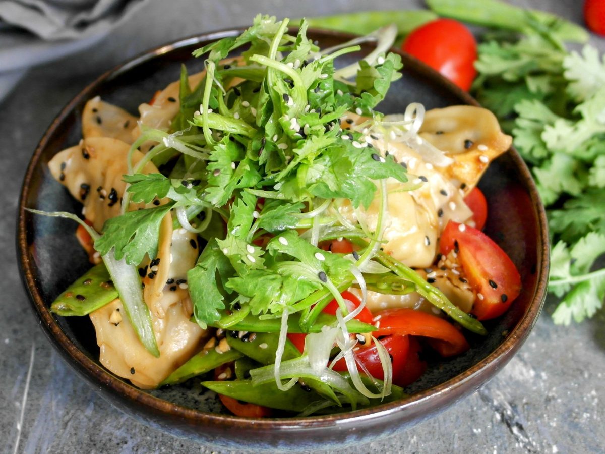 Eine schwarze Schüssel mitd em Gyoza-Nudelsalat udn Kräutern auf einem grauen Steintisch von schräg oben fotografiert.