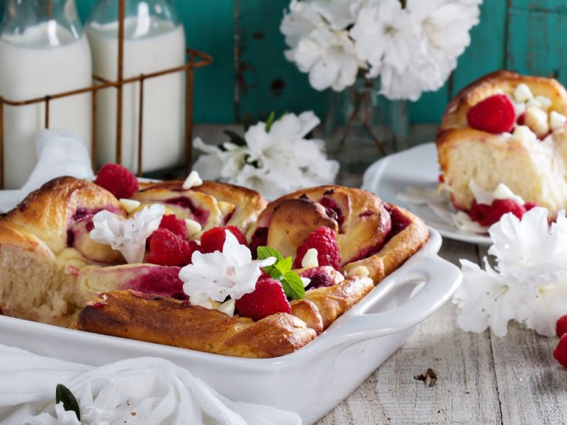 Frisch gebackene Himbeerschnecken mit frischen Himbeeren und weißer Schokolade in einer weißen Keramikform, dekoriert mit Blumen auf rustikalem Holztisch.