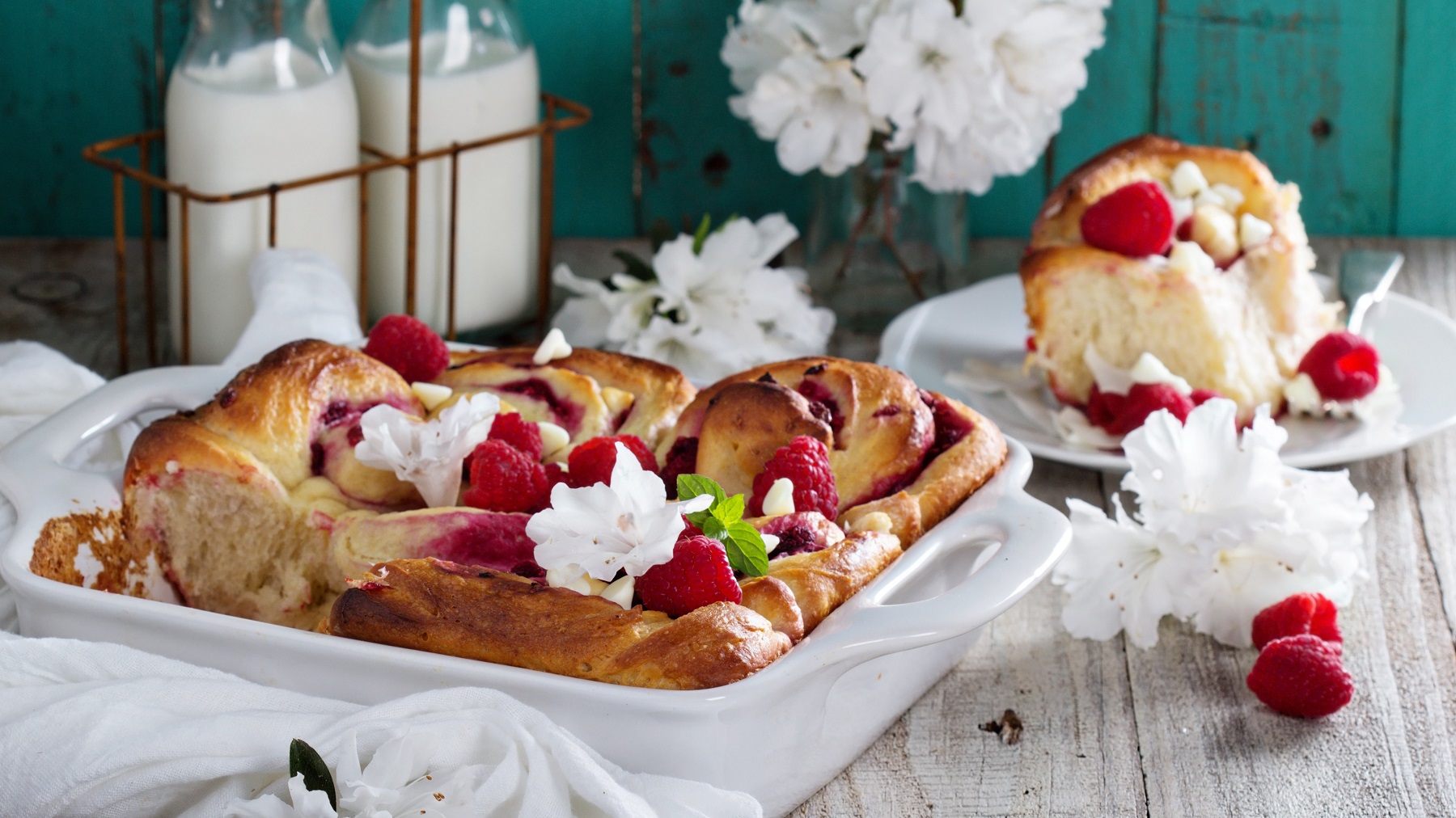 Frisch gebackene Himbeerschnecken mit frischen Himbeeren und weißer Schokolade in einer weißen Keramikform, dekoriert mit Blumen auf rustikalem Holztisch.