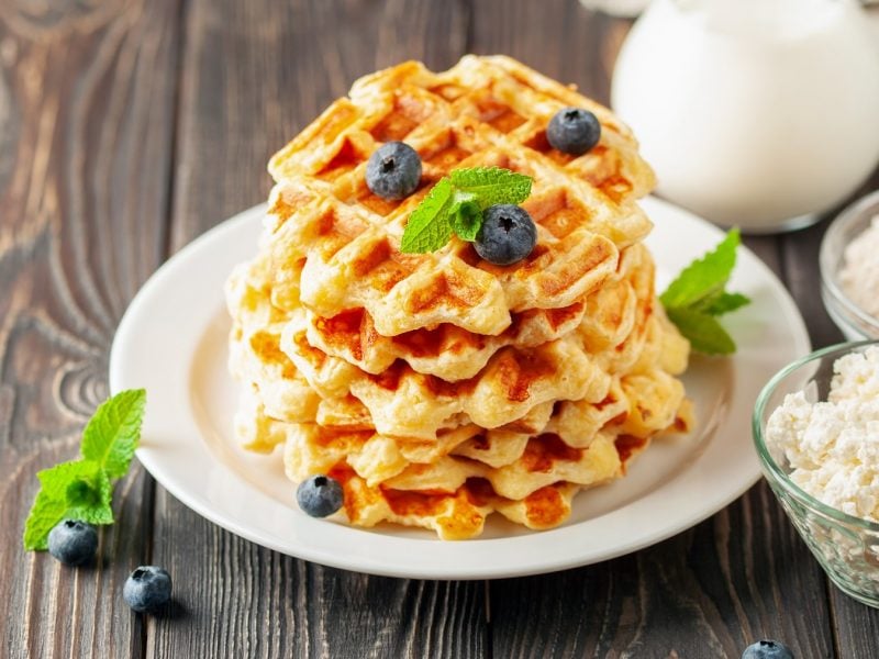 Mehrere Waffeln auf einem weißen Teller mit Blaubeeren und Minze auf einem rustikalen Holztisch.