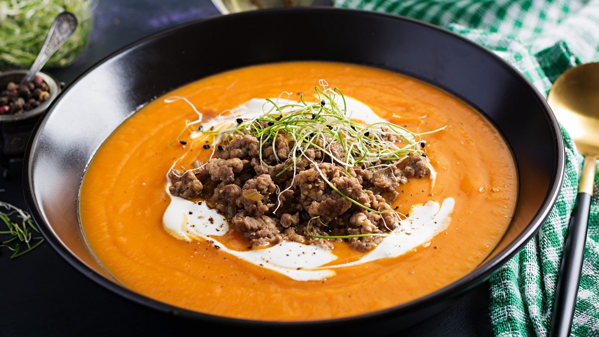 Die Low-Carb Cheeseburger-Suppe in einem schwarzen Teller auf dunklem Tisch mit grün-weiß-gestreiftem Küchentuch.