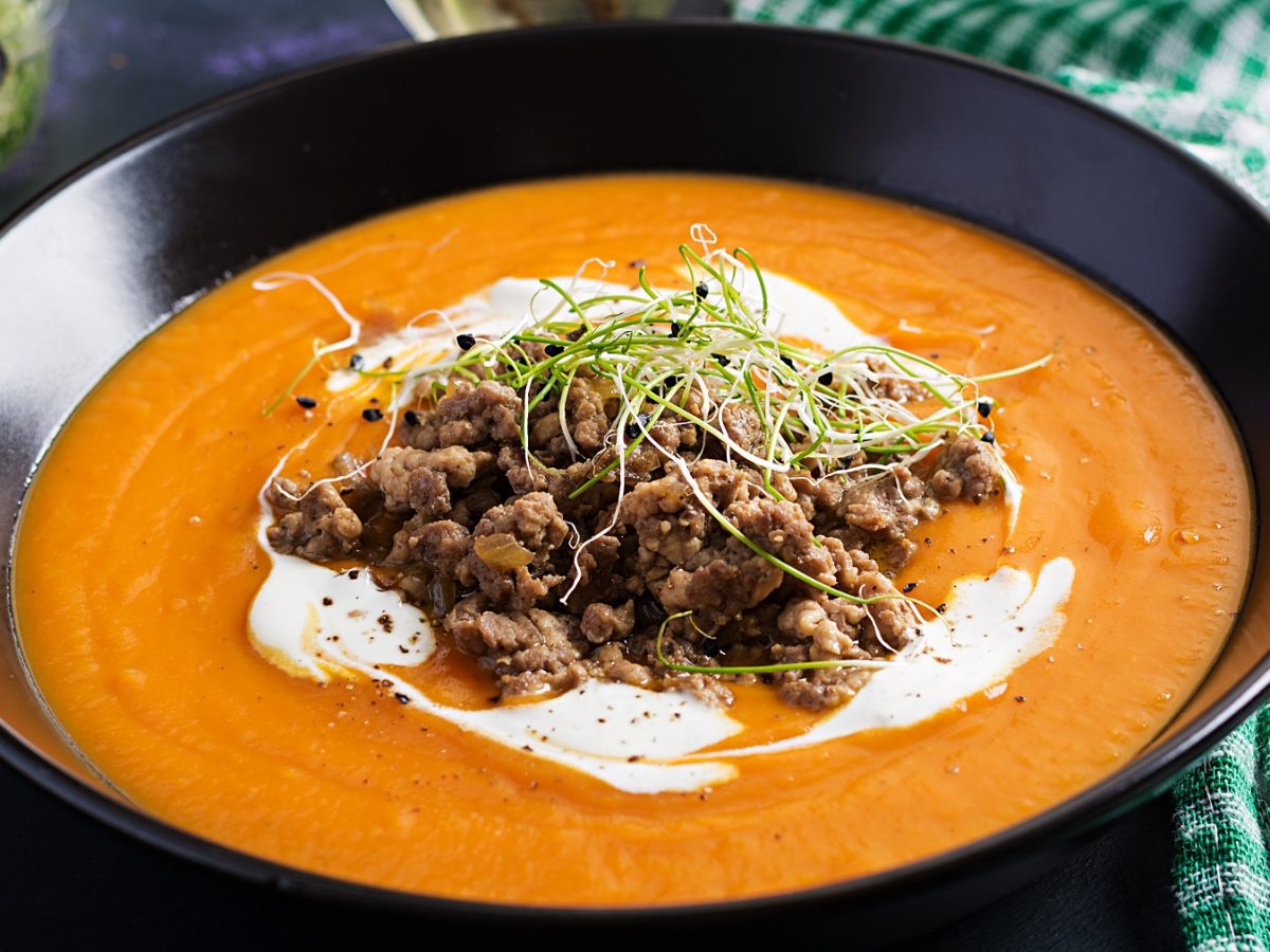 Die Low-Carb Cheeseburger-Suppe in einem schwarzen Teller auf dunklem Tisch mit grün-weiß-gestreiftem Küchentuch.