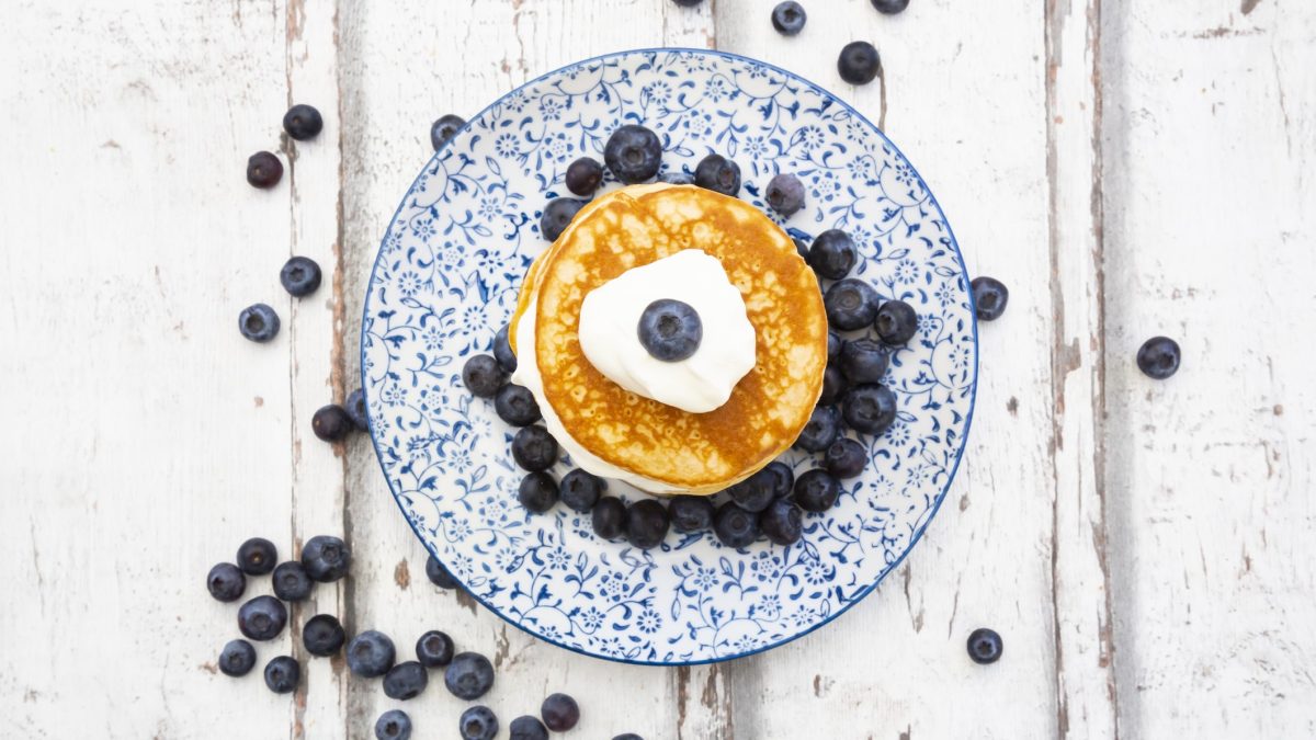 Ein blau-weißer Teller in klassischem Muster mit den Low-Carb-Pfannkuchen, Skyr und Blaubeeren auf einem hellen Holztisch. Alles von oben fotografiert.