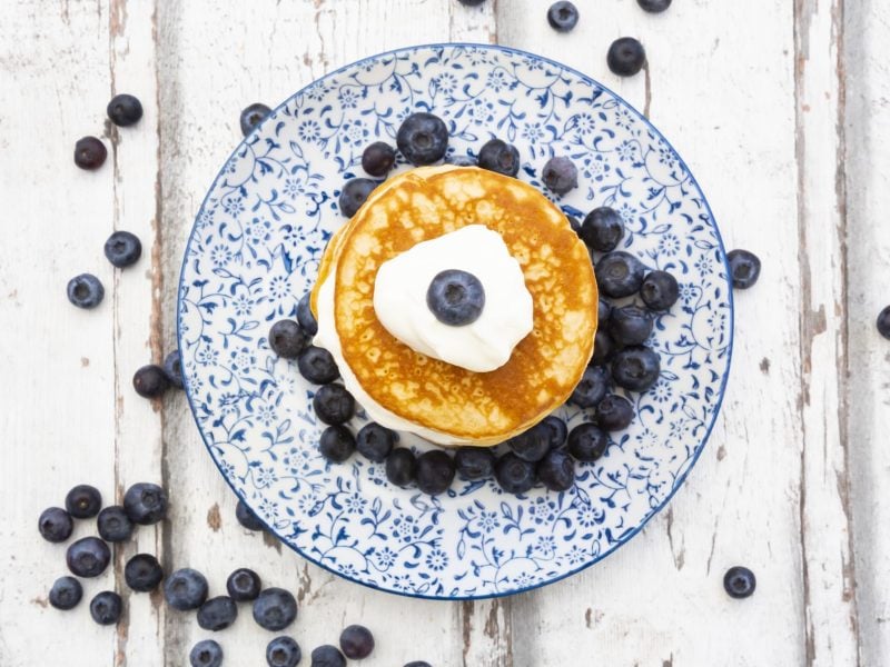 Ein blau-weißer Teller in klassischem Muster mit den Low-Carb-Pfannkuchen, Skyr und Blaubeeren auf einem hellen Holztisch. Alles von oben fotografiert.