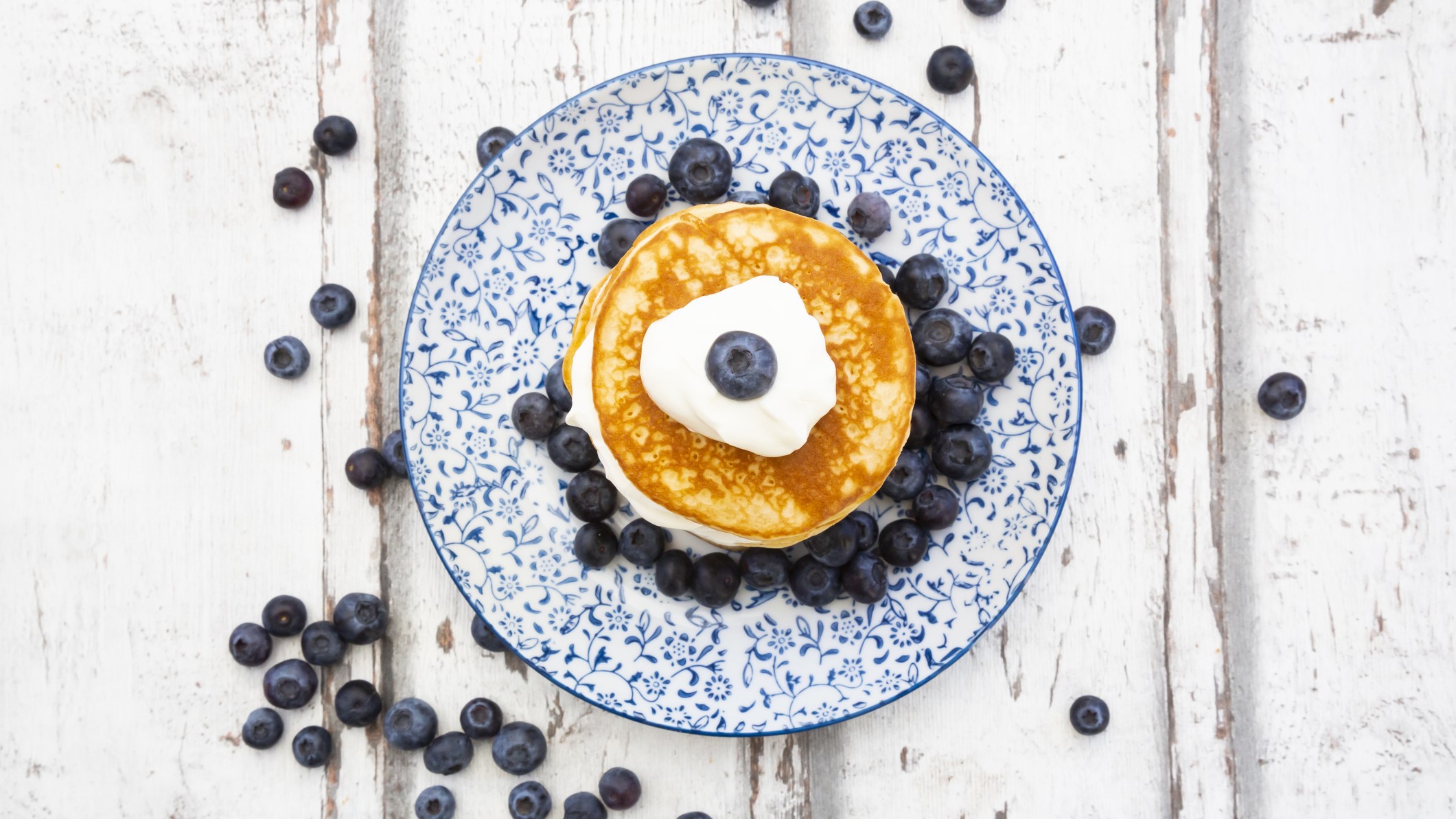 Ein blau-weißer Teller in klassischem Muster mit den Low-Carb-Pfannkuchen, Skyr und Blaubeeren auf einem hellen Holztisch. Alles von oben fotografiert.