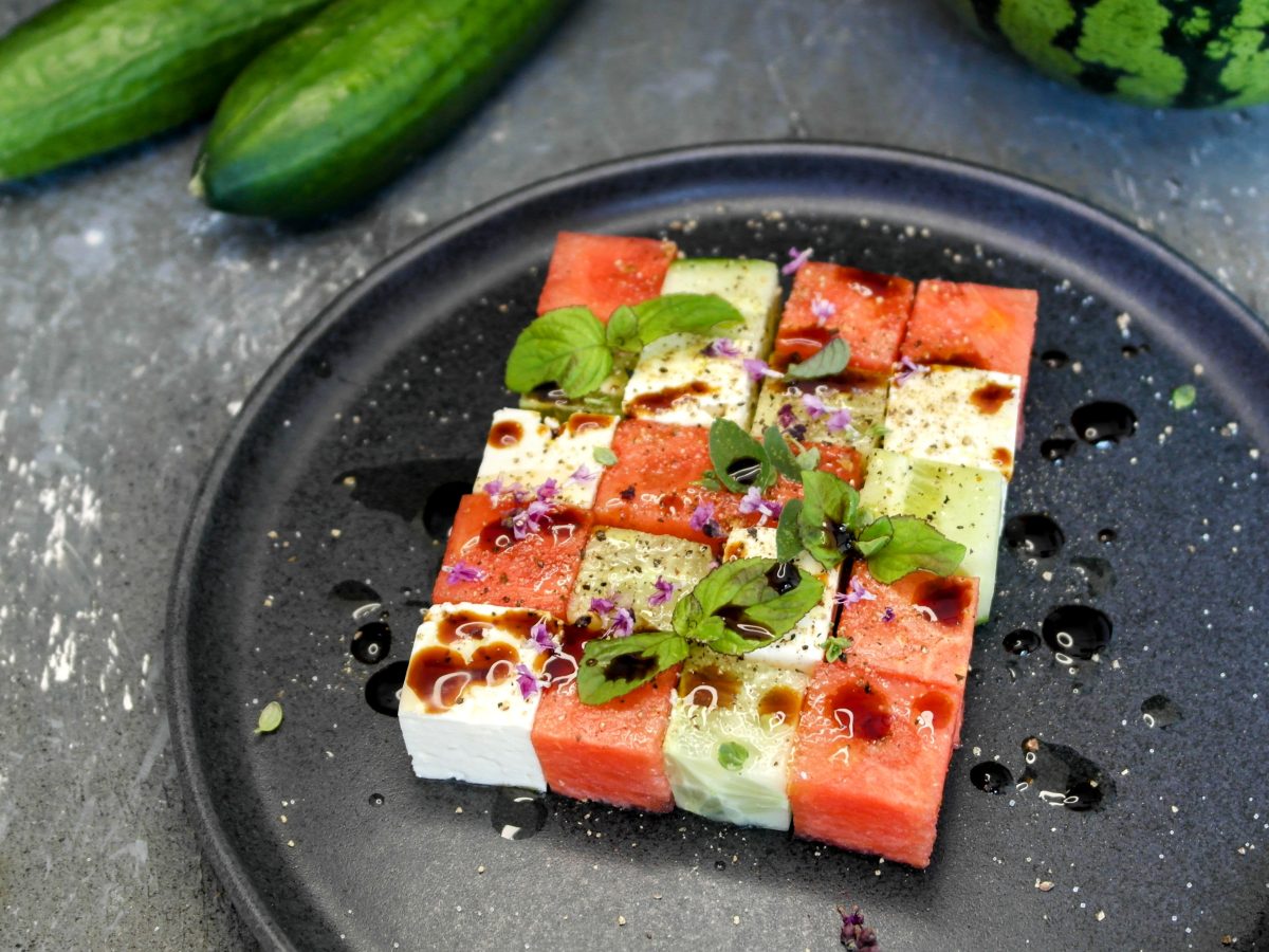 Ein grauer Teller mit dem gewürfelten Salat auf grauem Steintisch, mit Gurke und Wassermelone im Hintergrund. Alles von schräg oben fotografiert.