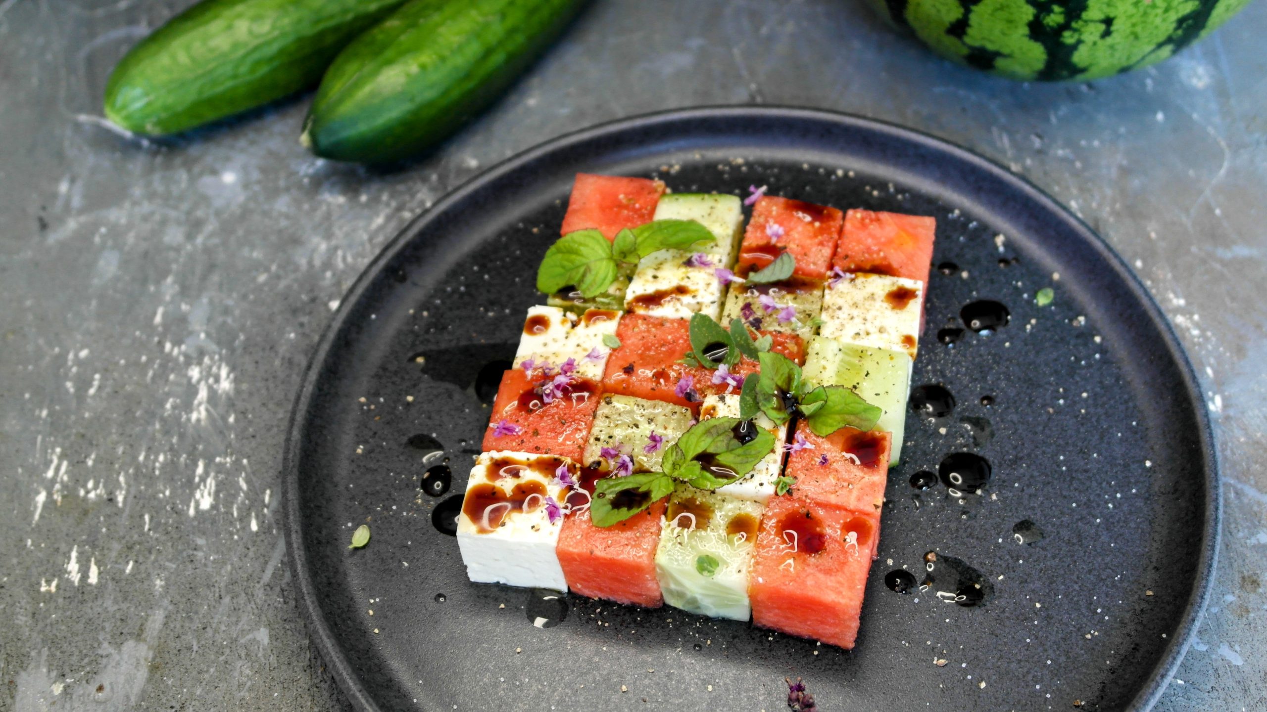 Ein grauer Teller mit dem gewürfelten Salat auf grauem Steintisch, mit Gurke und Wassermelone im Hintergrund. Alles von schräg oben fotografiert.