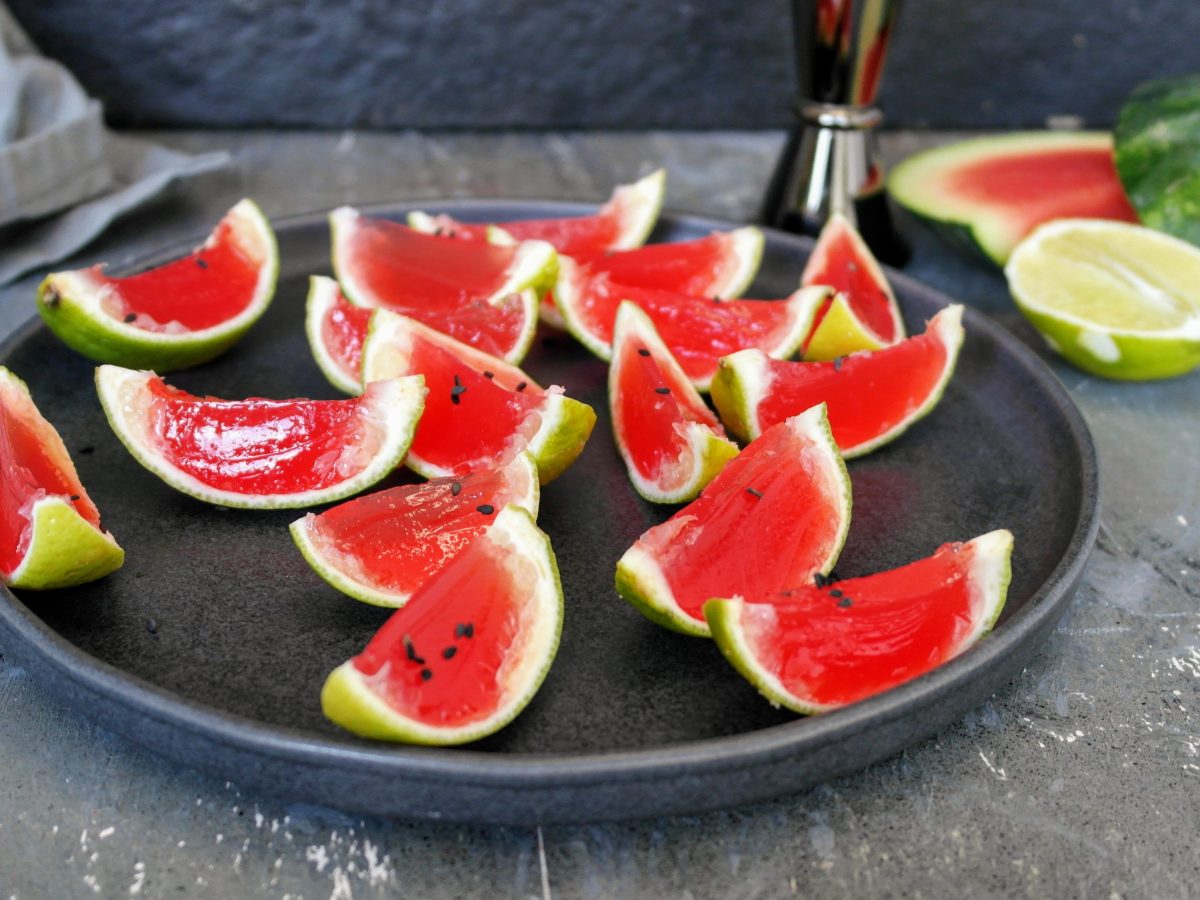 Ein schwarzer Teller mit vielen Spalten der Mini-Wassermelone mit Tequila auf grauem Tisch mit schwarzem Hintergrund. Alles von leicht schräg oben fotografiert.