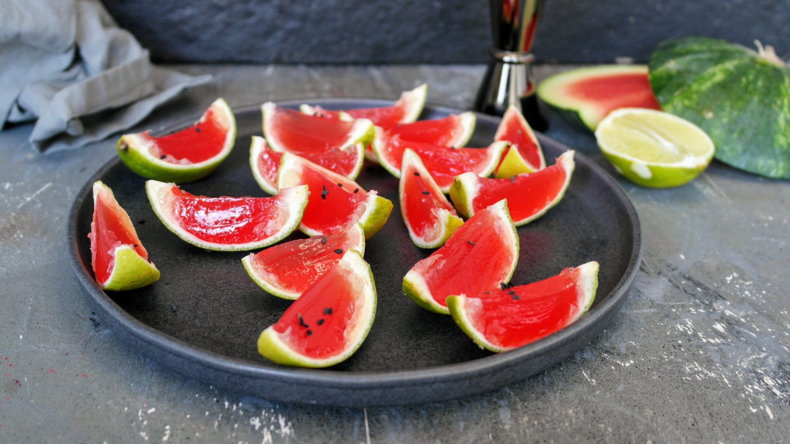 Ein schwarzer Teller mit vielen Spalten der Mini-Wassermelone mit Tequila auf grauem Tisch mit schwarzem Hintergrund. Alles von leicht schräg oben fotografiert.