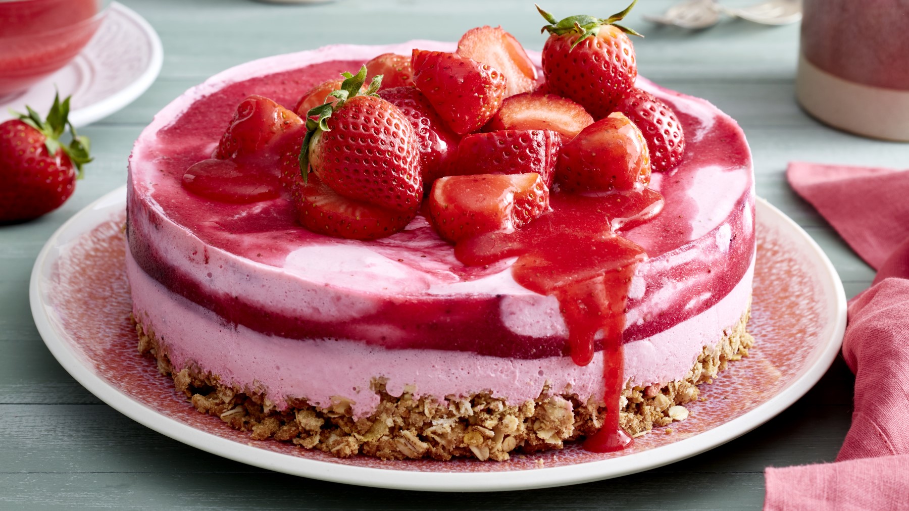 Müsli-Erdbeertorte mit frischen Erdbeeren und Sauce darauf auf einem rosa Teller.