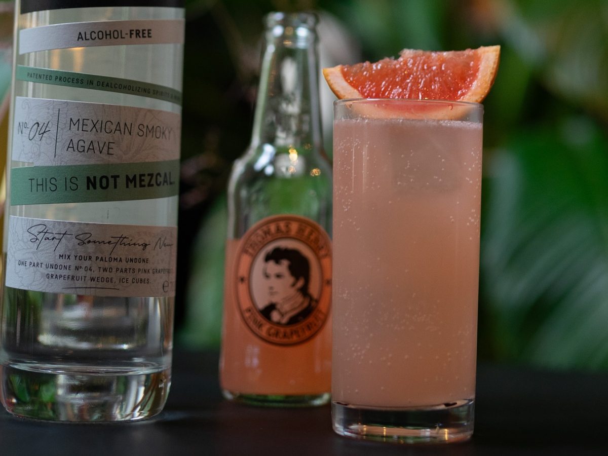 Paloma Undone in einem Highballglass mit Grapefruitschnitz, daneben Flaschen von Undone und Thomas Henry.