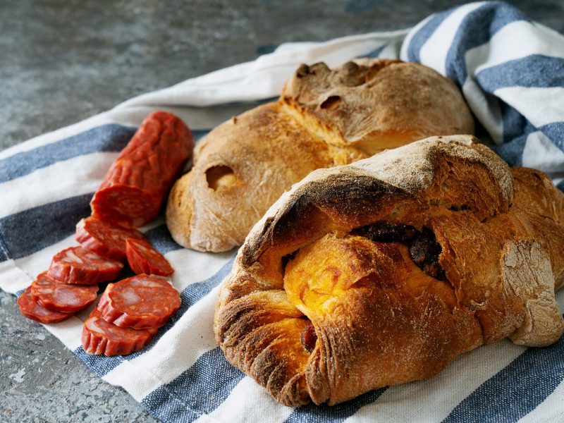 Zwei rustikale Pão com Chouriço und aufgeschnittene Chouriço liegen auf einem blau-weiß gestreiften Tuch auf grauem Steinboden.