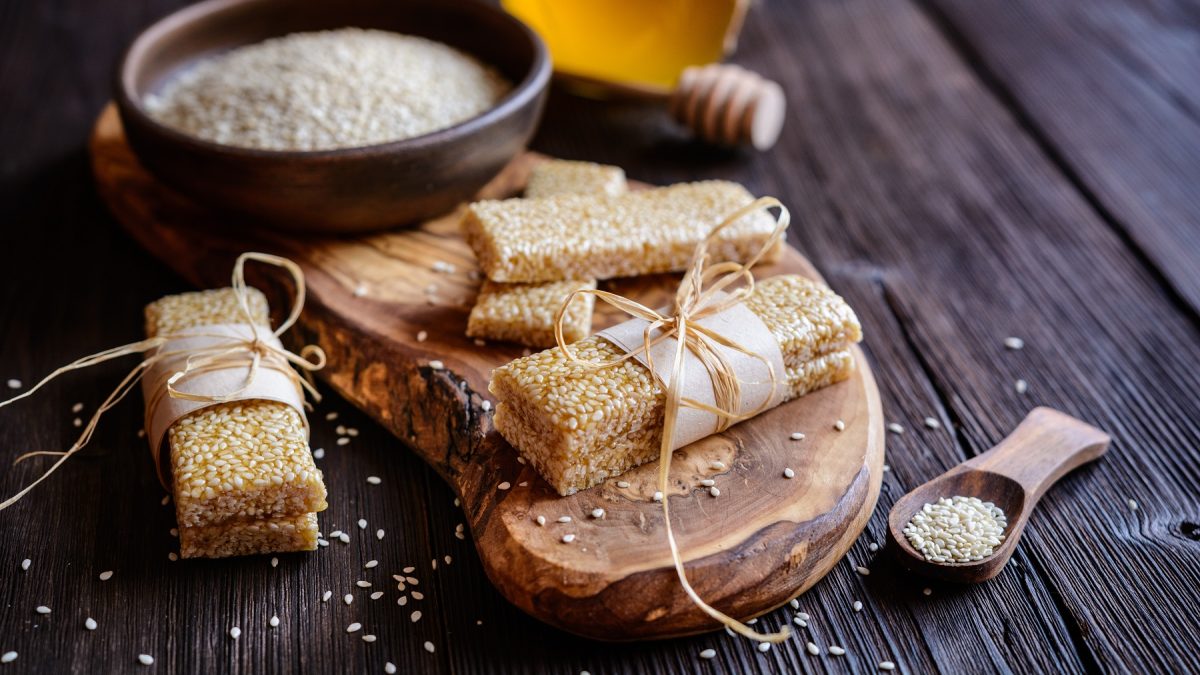 Pasteli mit Band auf Holzbrett neben einer Schale Sesam und Honiglöffel auf dunklem Holztisch arrangiert.