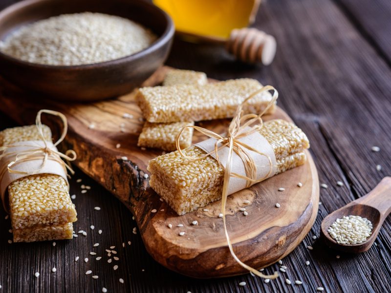 Pasteli mit Band auf Holzbrett neben einer Schale Sesam und Honiglöffel auf dunklem Holztisch arrangiert.