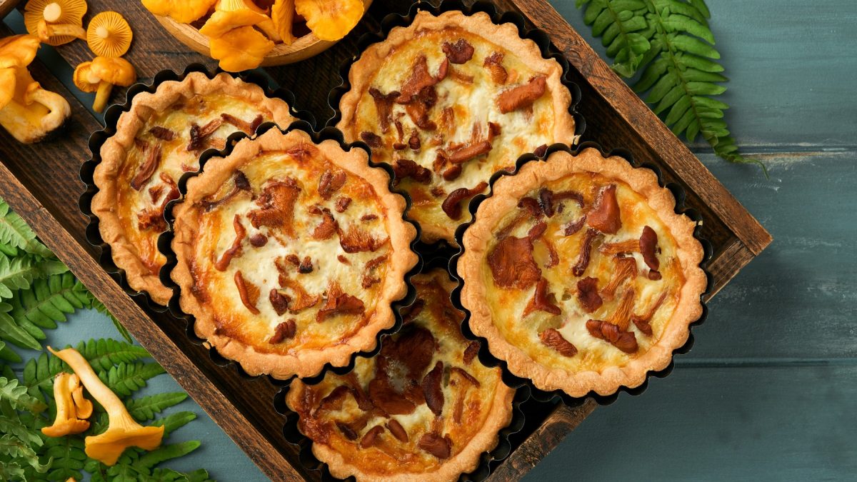 Mehrere Pfifferling-Tartelettes auf einem dunklen Schneidebrett und dunkelgrünem Tisch. Alles von oben fotografiert.