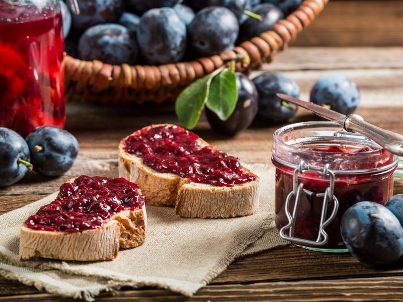 Zwei Scheiben Brot mit Pflaumenmarmelade, daneben ein Glas Marmelade und frische Pflaumen auf rustikalem Holztisch.