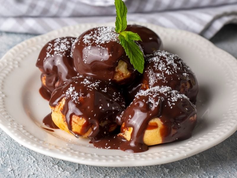 Ein weißer Teller mit den Profiterole und Schokosauce auf einem grauen Tisch mit grau-weiß-gestreifter Tischdecke.