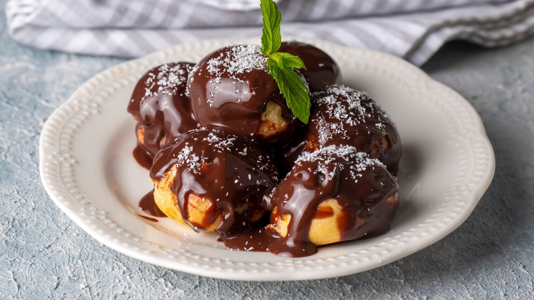 Ein weißer Teller mit den Profiterole und Schokosauce auf einem grauen Tisch mit grau-weiß-gestreifter Tischdecke.