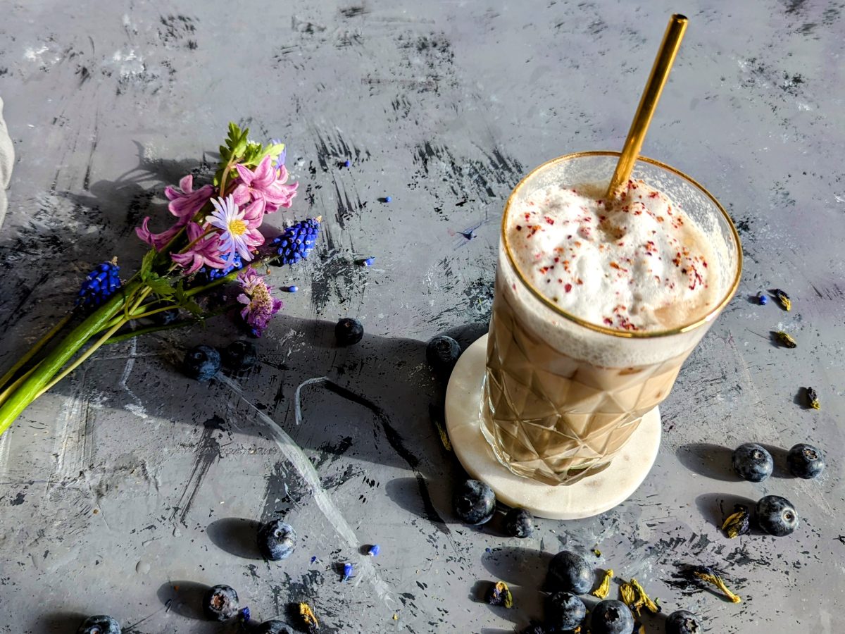 Ein Glas mit dem Blaubeer-Latte auf grauem Steintisch in der Sonne mit Blüten und Blaubeeren darauf verteilt. Alles von schräg oben fotografiert.