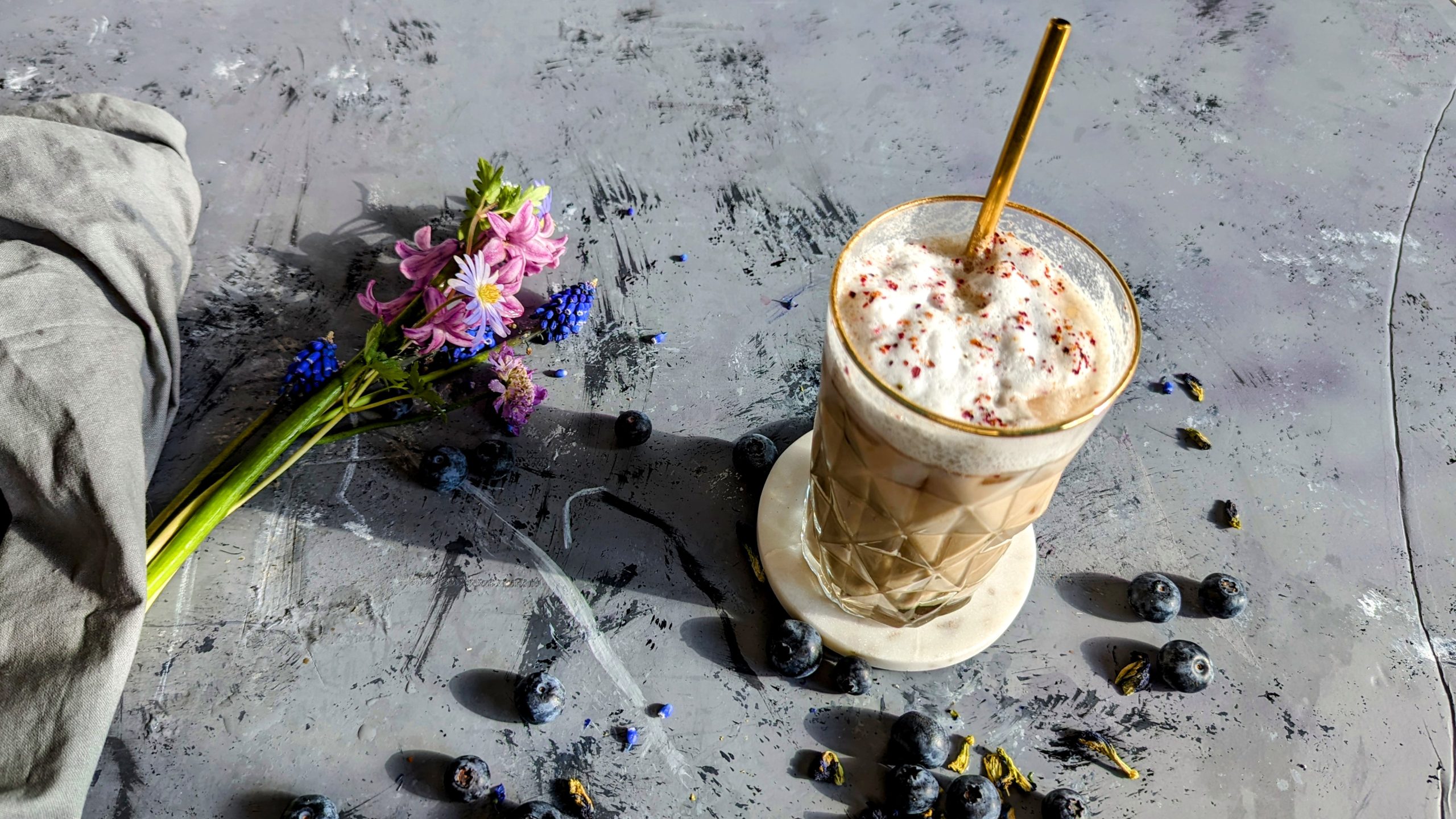 Ein Glas mit dem Blaubeer-Latte auf grauem Steintisch in der Sonne mit Blüten und Blaubeeren darauf verteilt. Alles von schräg oben fotografiert.